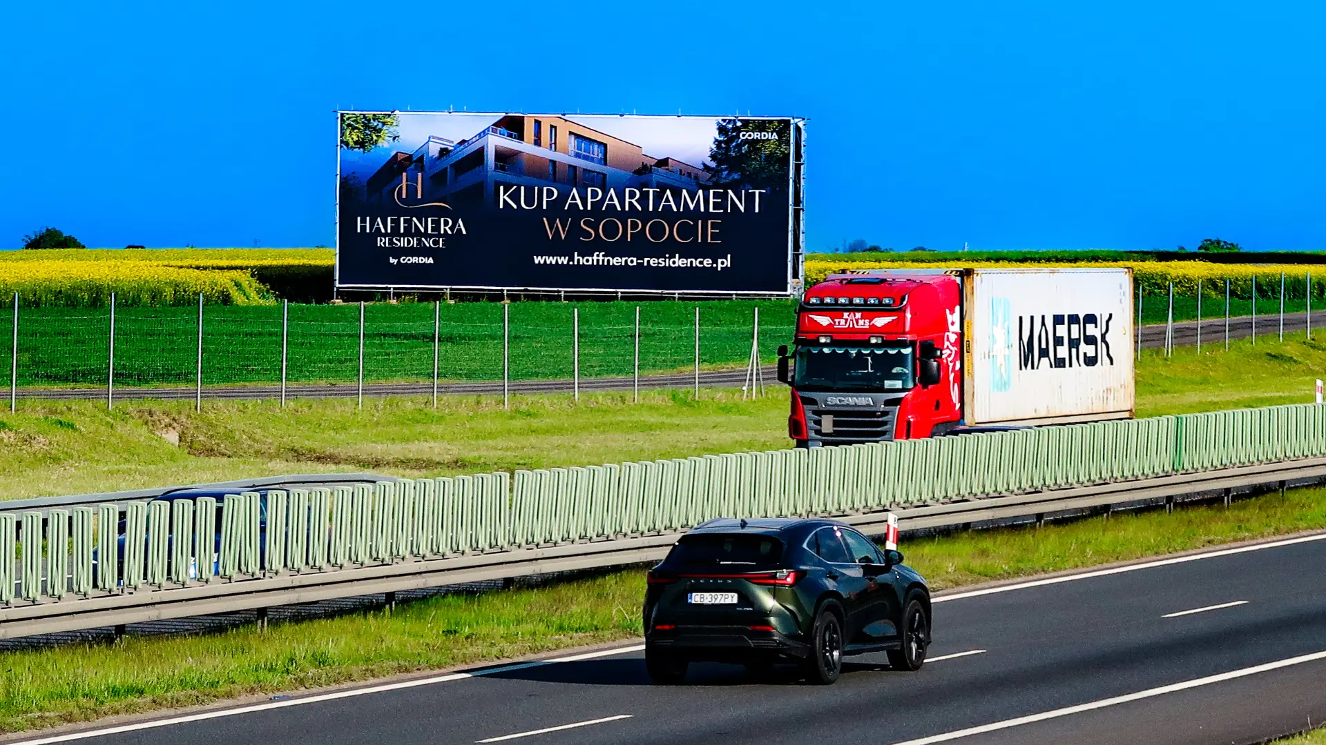 Wielkoformatowy nośnik reklamowy z promocją apartamentów Haffnera Residence, zlokalizowany przy trasie, widoczny za przejeżdżającą ciężarówką i samochodem osobowym.