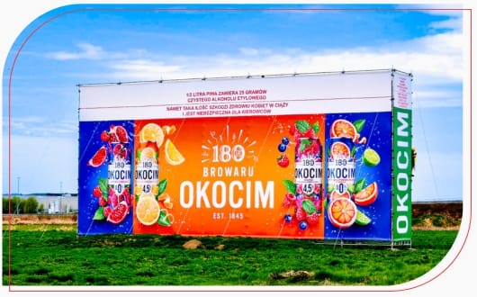 Wielkoformatowy nośnik reklamowy Okocim na tle nieba, ustawiony w terenie zielonym, przy trasie szybkiego ruchu
