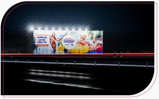 Podświetlony nocą wielkoformatowy nośnik reklamowy Suntago przy trasie szybkiego ruchu, promujący wakacje przez cały rok