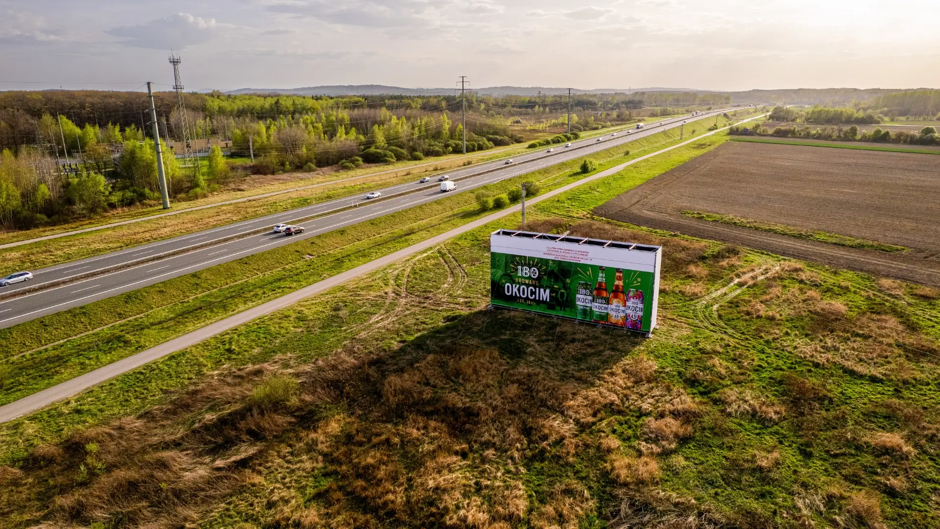 Reklama Okocim na dużym nośniku w otwartym terenie, widoczna z jadących samochodów przy trasie szybkiego ruchu