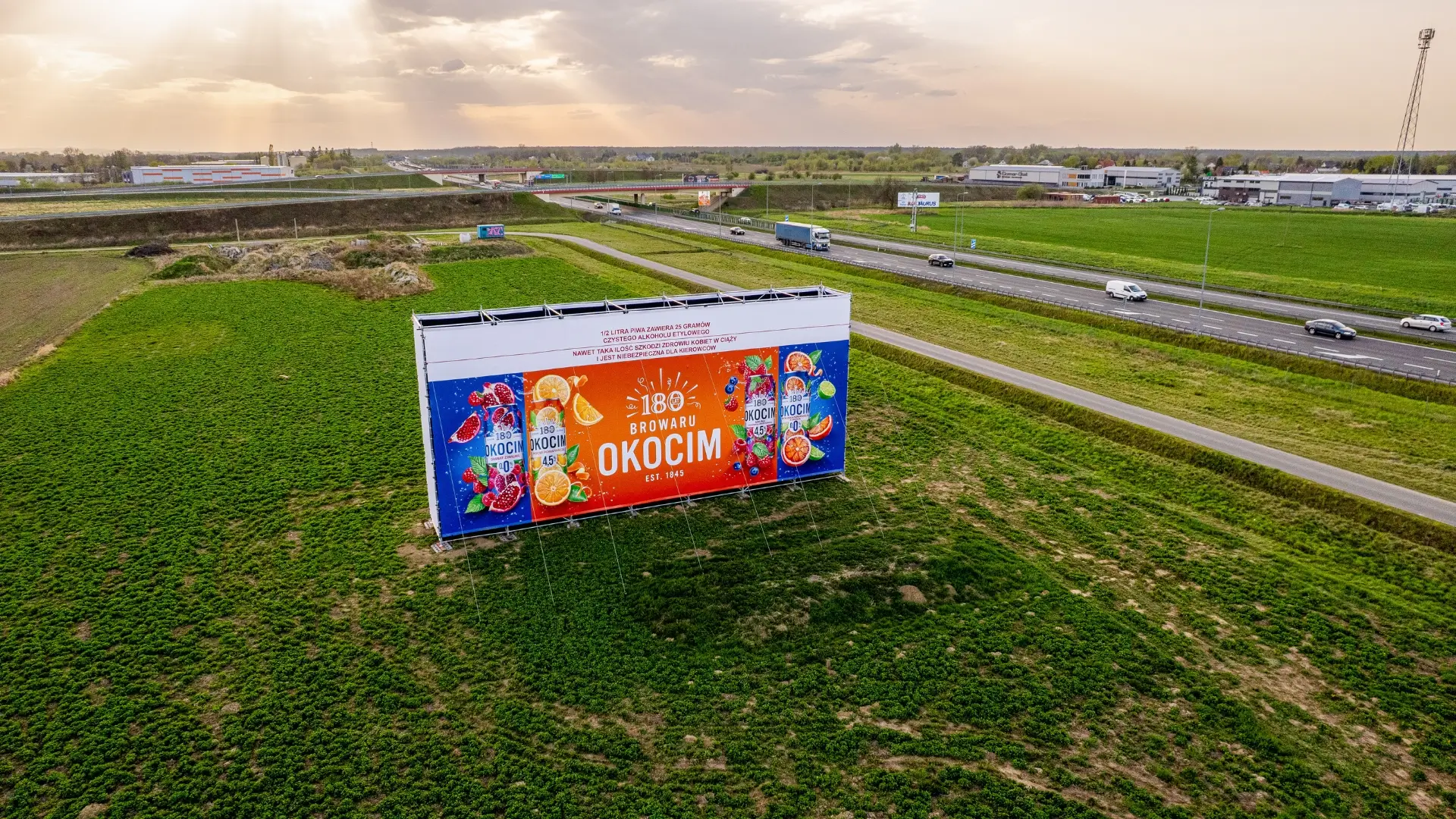 Wielkoformatowy billboard marki Okocim zlokalizowany przy ruchliwej trasie, widziany z perspektywy lotu dronem, na tle zielonego terenu i wieczornego nieba.