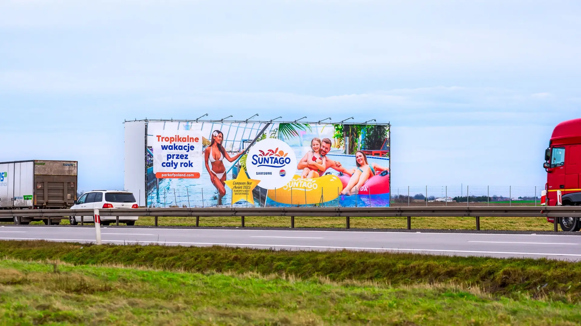 Wielkoformatowa reklama Suntago widoczna z drogi ekspresowej, zamontowana na konstrukcji w otwartym terenie, obok przejeżdżających pojazdów.